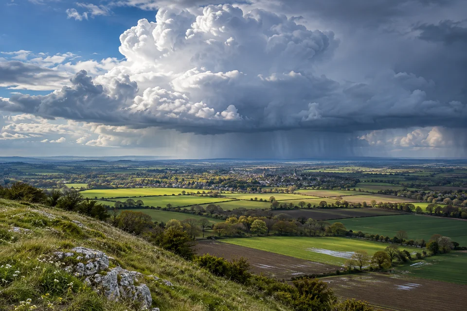 A towering rain cloud and distant rain shafts pass over imagined fields and a town.