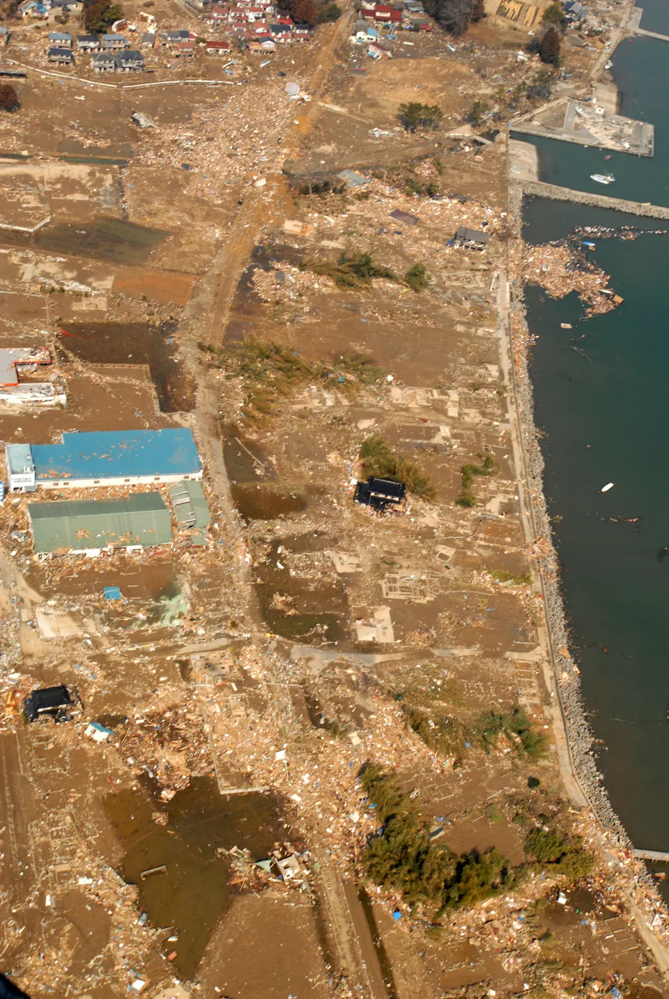 Tsunami damage at Kawara fishing port, Kesennuma, photographed from a U.S. Navy helicopter on 13 March 2011. Debris covers the coastal plain beside the harbour.